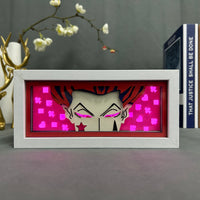 Boite lumineuse Hisoka cadre blanc posée sur bureau, sourire caractéristique et yeux roses sur fond fuchsia avec cartes