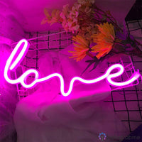 Néon LED inscription love en cursive rose fuchsia posé sur tissu blanc avec fleurs colorées