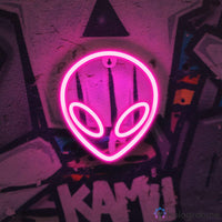Néon LED alien rose uni en forme de tête extraterrestre avec yeux ovales accroché sur mur graffiti, déco gaming