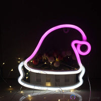 Néon LED bonnet de Noël rose et blanc lumineux posé près de fleurs et guirlandes dorées