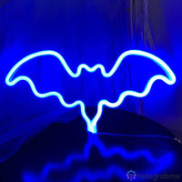 Néon LED chauve-souris bleu ciel aux ailes ouvertes posé sur socle, lumière vive sur fond bleu