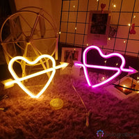 Deux néons LED cœur cupidon jaune et rose avec flèche posés côte à côte sur tapis moelleux