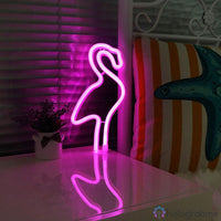 Néon LED flamant rose rose vif posé sur une table de nuit, ambiance chambre féminine avec coussins