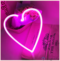 Néon LED grand cœur rose fuchsia lumineux tenu devant des fleurs roses, décoration romantique