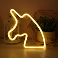 Néon LED licorne blanc chaud posé sur un bureau entre une lanterne et un pot de plante, ambiance chambre