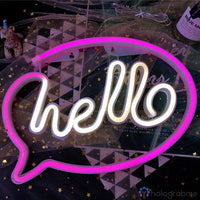 Néon LED hello en cursive blanche dans une bulle de dialogue rose, posé sur table avec confettis
